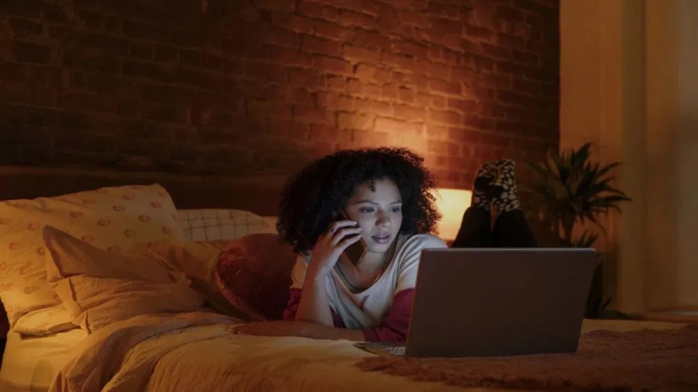 Une femme allongée sur son lit parlant sur son téléphone tout en utilisant un ordinateur portable.